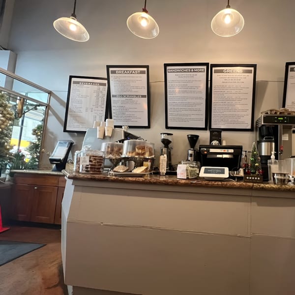 Cafe La Tazita's counter with pastries, coffee machines, and menu boards for breakfast, sandwiches, and greens.