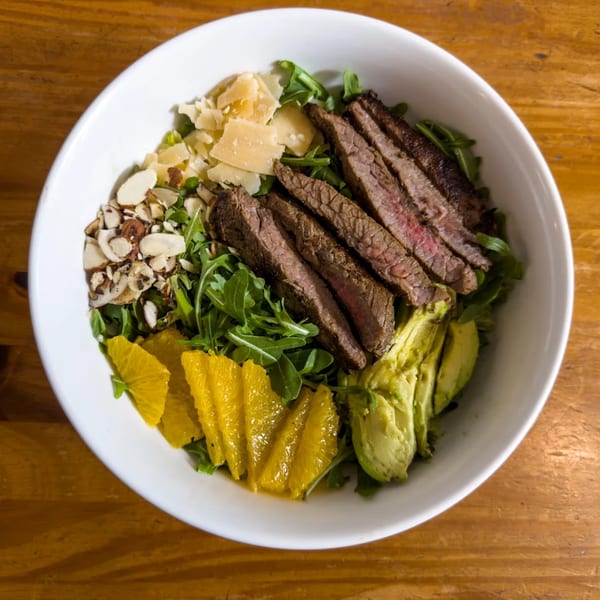 Steak salad with arugula, avocado, orange slices, almonds, and parmesan cheese in a white bowl.