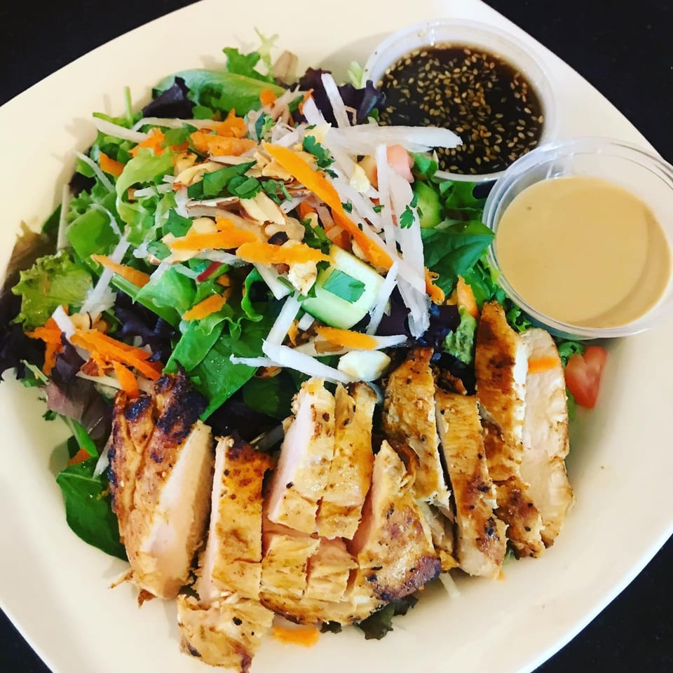 Grilled chicken salad with mixed greens, shredded carrots, and two dressing cups on a white plate.